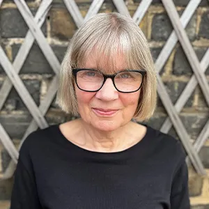 Dr Rachel Baggaley OBE with short, straight grey hair and black glasses, smiling softly. She is wearing a black top and she is posed in front of a textured stone wall.