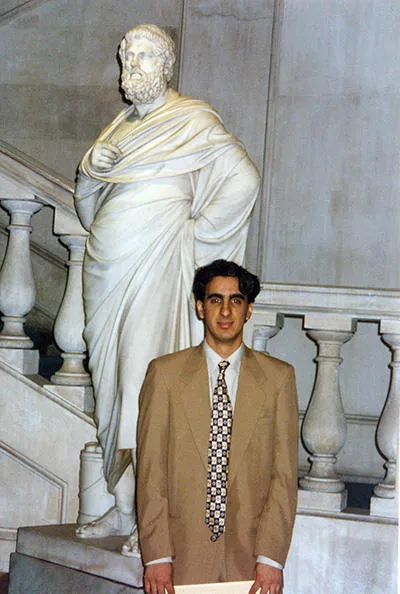 A young man with dark hair is wearing a light brown suit with a shirt and tie. He is standing next to a marble statue in front of the main staircase at the King’s Building on the Strand. 