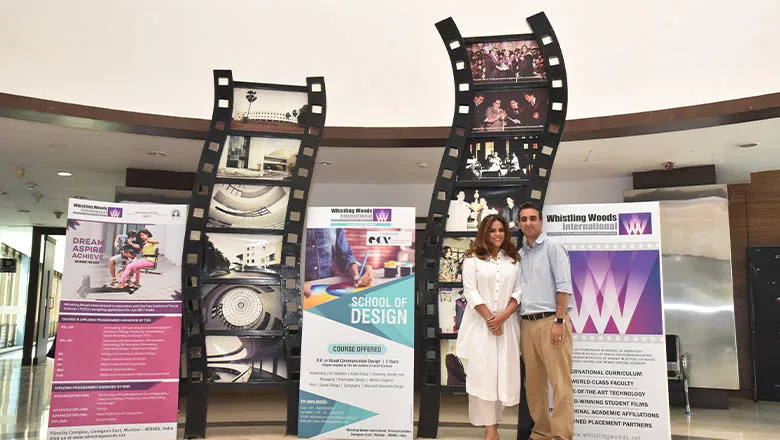 A man and woman proudly stand at the stylish entrance to a chic-looking building. Behind them are boards displaying the courses and achievements of Whistling Woods International 
