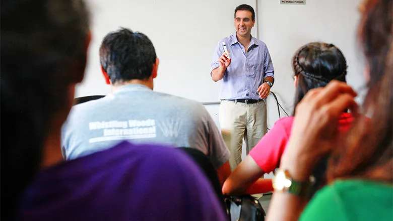 A man with dark hair and wearing grey trousers and a blue shirt stands before whiteboard. He is enthusiastically talking to a classroom of students. 