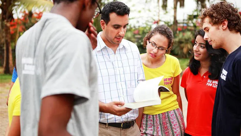 A man with dark hair who is wearing a checked white shirt stands in the middle of several students. He is reading a book and explaining something to the group. 