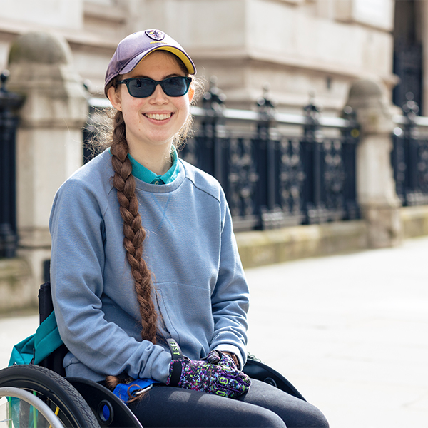 Rebecca, with a long braid, smiling as she sits in a wheelchair outdoors, wearing a light blue sweatshirt, patterned gloves, dark sunglasses, and a baseball cap. A stone building and black railings are visible in the background.