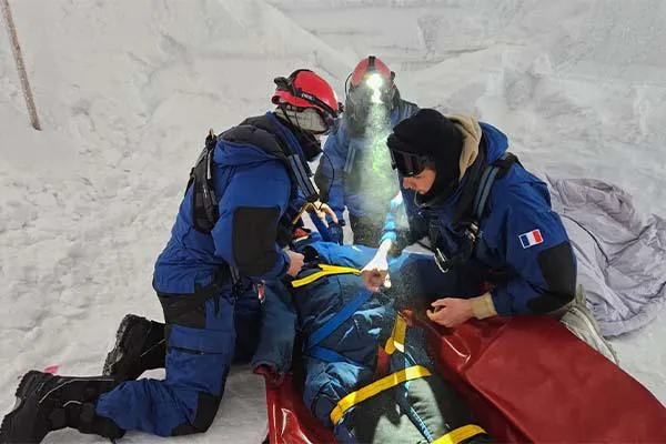 Three individuals in winter gear are engaged in a rescue exercise on a snowy surface. They are assisting a simulated victim, who is lying on a stretcher. 