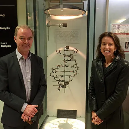 A woman with long brown hair and wearing a black leather jacket stands next to a display cabinet. This houses a scientific model showing the structure of DNA. To her right is a man wearing a dark grey suit. 