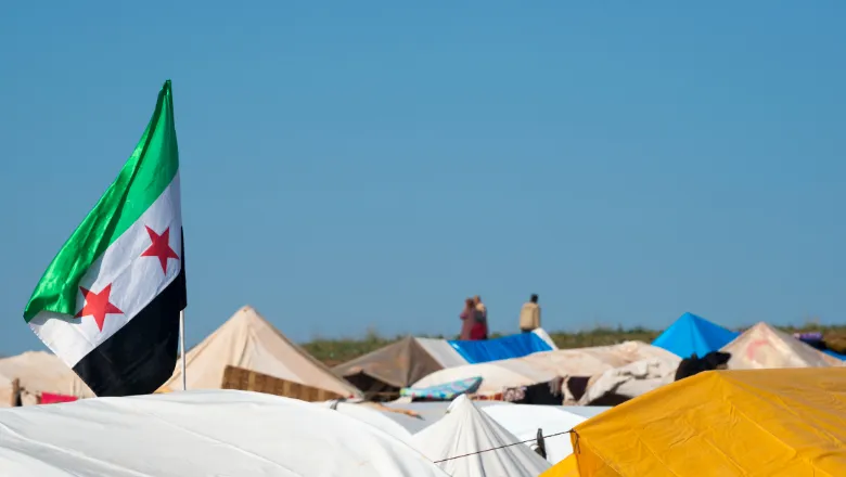 A picture showing the tops of tents in a refuee camp. Two people can be seen in the distance. A syrian flag waves prominently in the foreground.