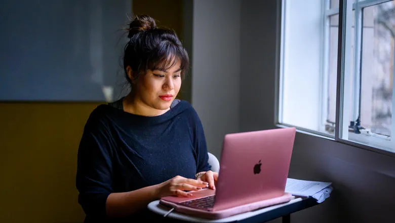 A woman sits with her laptop by a window.