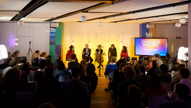 5 panellists in front of yellow-orange lighting and audience with a screen showing 