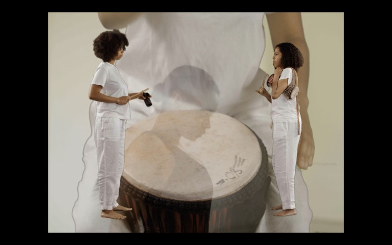 Two Black women in white on either side of a drum on which there is the shadow of a third woman