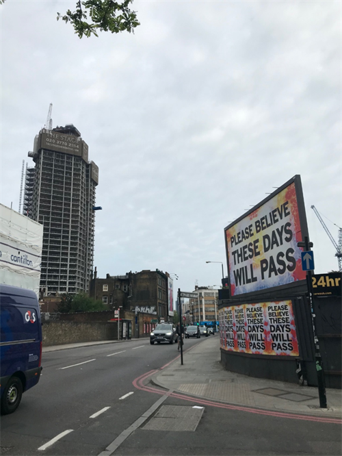 Photo of a road in Shoredich. A billboard has a large colourful sign which says "please believe these days will pass".
