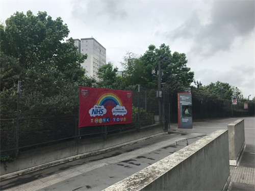 Photo of padestrian walkway at Emirates Stadium, Holloway with a large red banner attached to the railings. The banner has arsenal logos on it, a rainbow and the text reads "To our NHS and all the Key Workers, thank you"