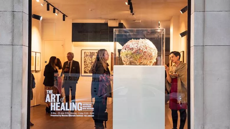 Front window of The Art of Healing exhibition, featuring two people looking at a plinth displaying a nest-like artwork by Ilawanti Ken