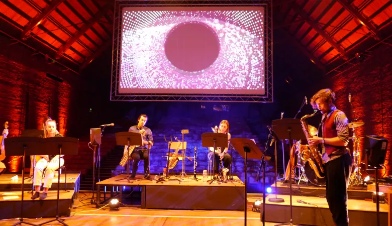 Christo playing the saxophone on a stage with other musicians in front of a screen visualising cosmic rays data