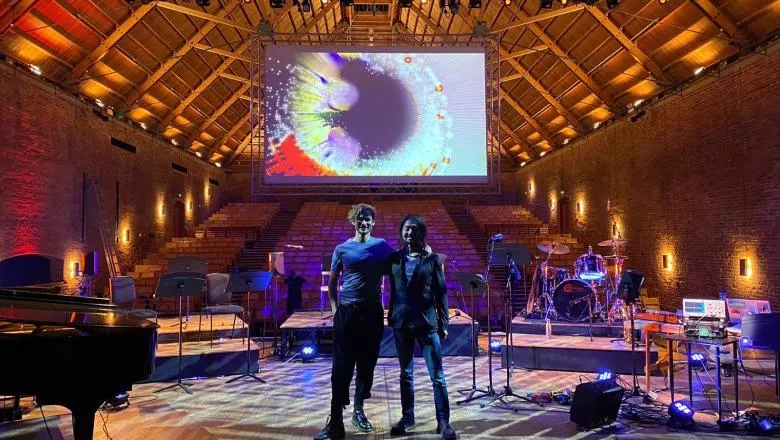Christo and Teppei posing  in front of stage with screen displaying visualised cosmic ray data