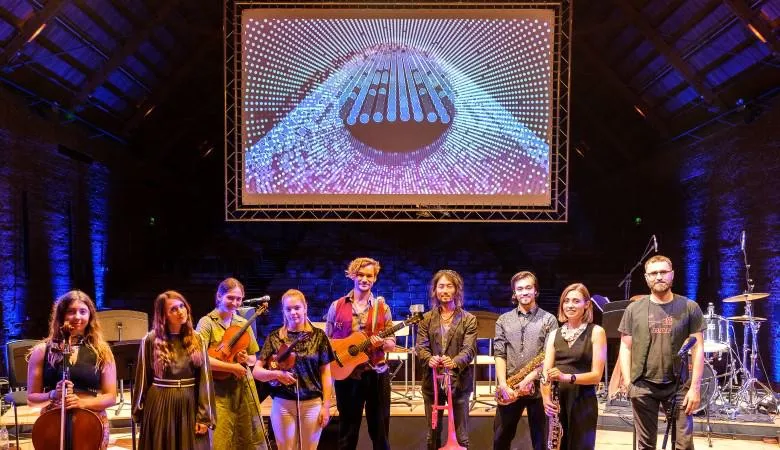 All musicians posing with their instruments and with Christo and Teppei in front of stage with screen visualising cosmic ray data