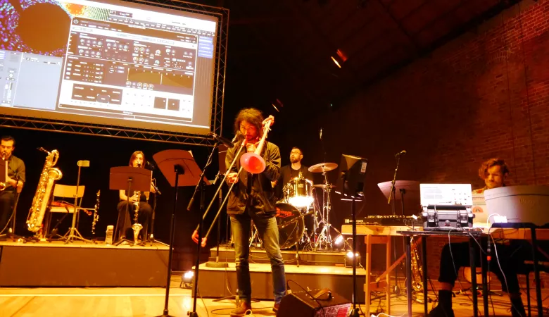 Teppei playing the trombone on a stage with other musicians in front of a screen visualising cosmic rays data