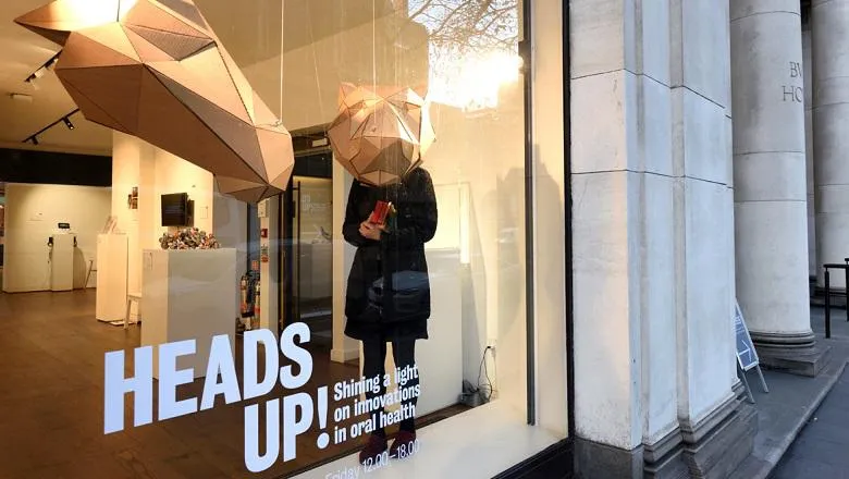 An image of the Arcade front window displaying the exhibition title and two, large cardboard animal heads with a visitor looking into one of the heads