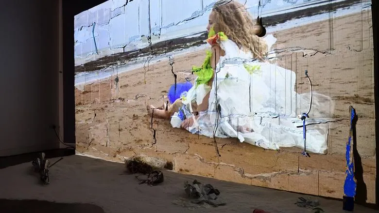 A photo of a large projection screen showing a film with a young girl sitting on the beach. There is sand on the floor in front of the screen with found objects featured in the film.