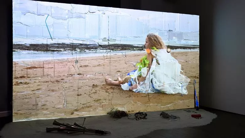 Image showing a projection screen with film still of a young woman sat on the beach. There is sand in front of the screen with objects used in the film.