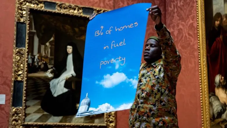 Black man inside a gallery holding a poster with the words '13% of homes in fuel poverty' against a blue sky background
