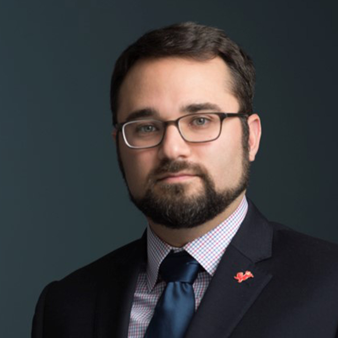 Dan Elinoff, a person with dark short hair and a full beard, dressed in a dark blazer, white shirt and blue tie.