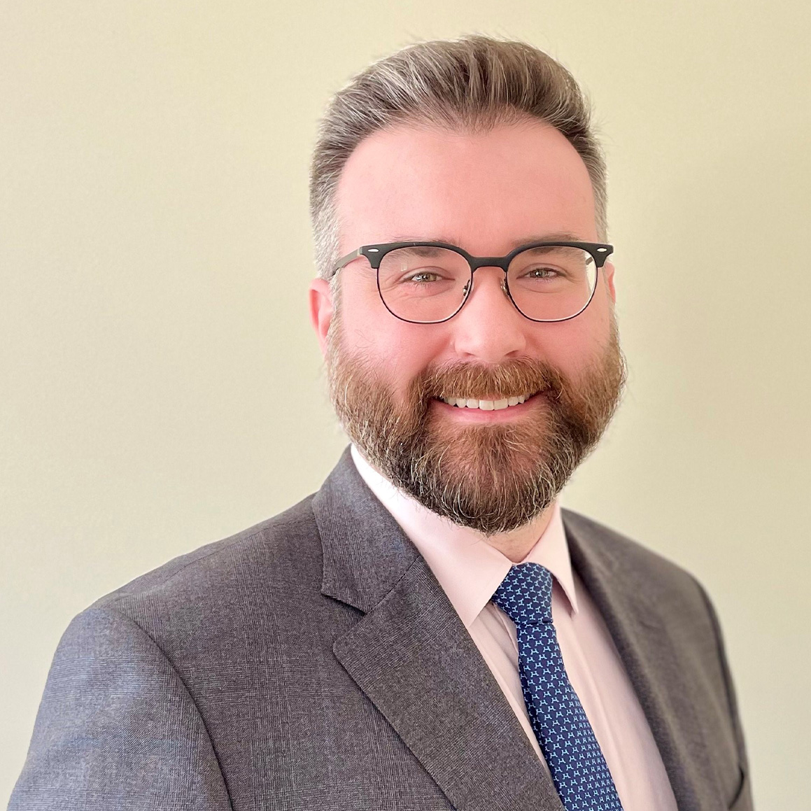 John F.Higgins with short brown hair and a full beard, dressed in a grey blazer, white shirt and a blue tie, smiling warmly.