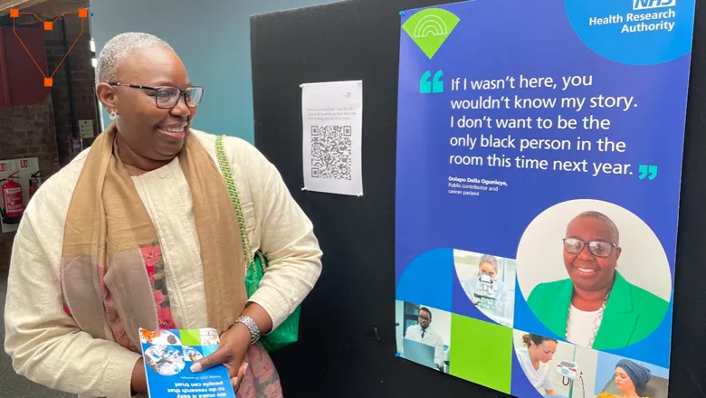 A woman (della), standing to the right of an NHS poster and smiling