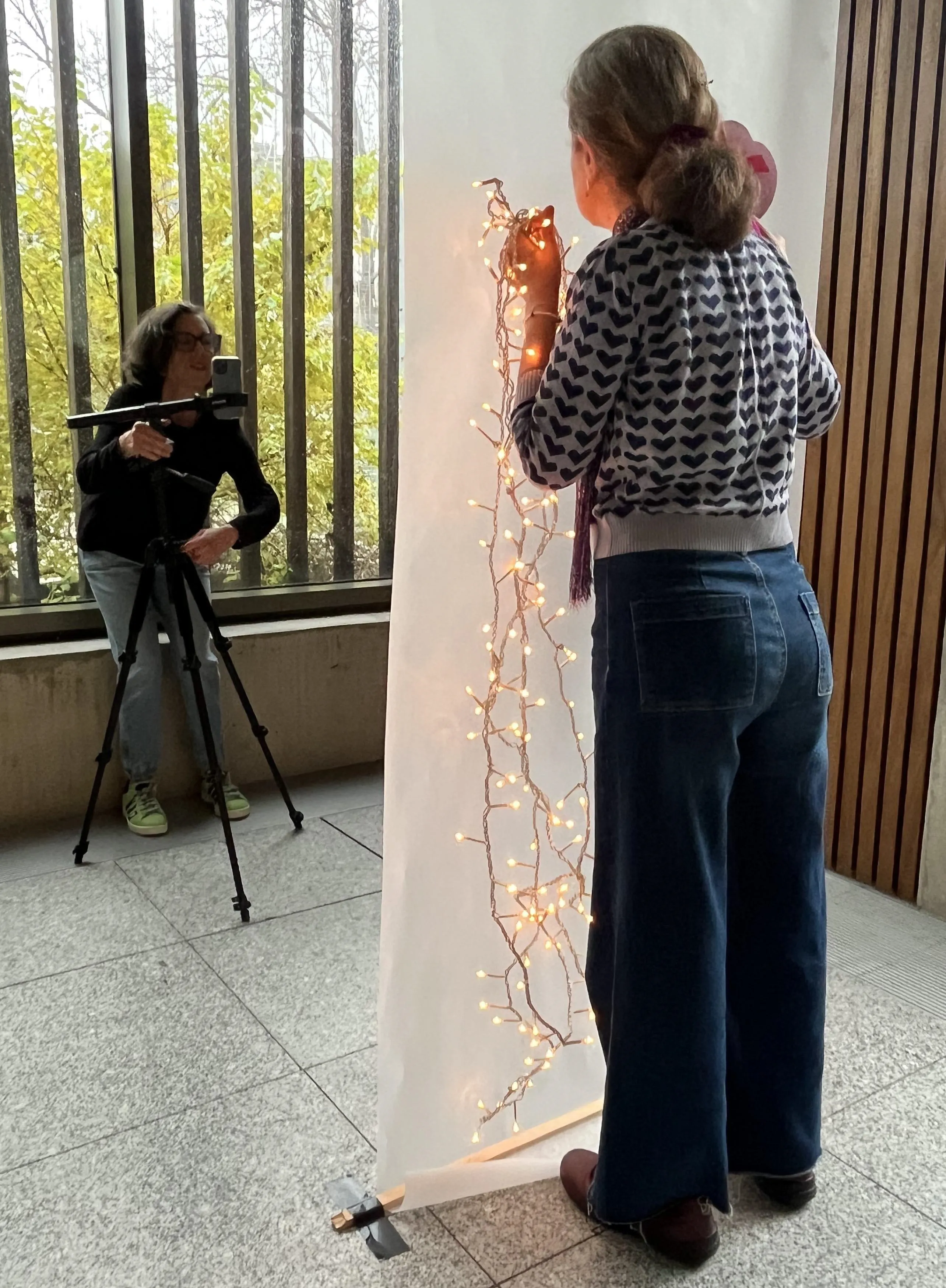 woman standing behind screen with fairy lights and another woman taking her photo with a smart phone