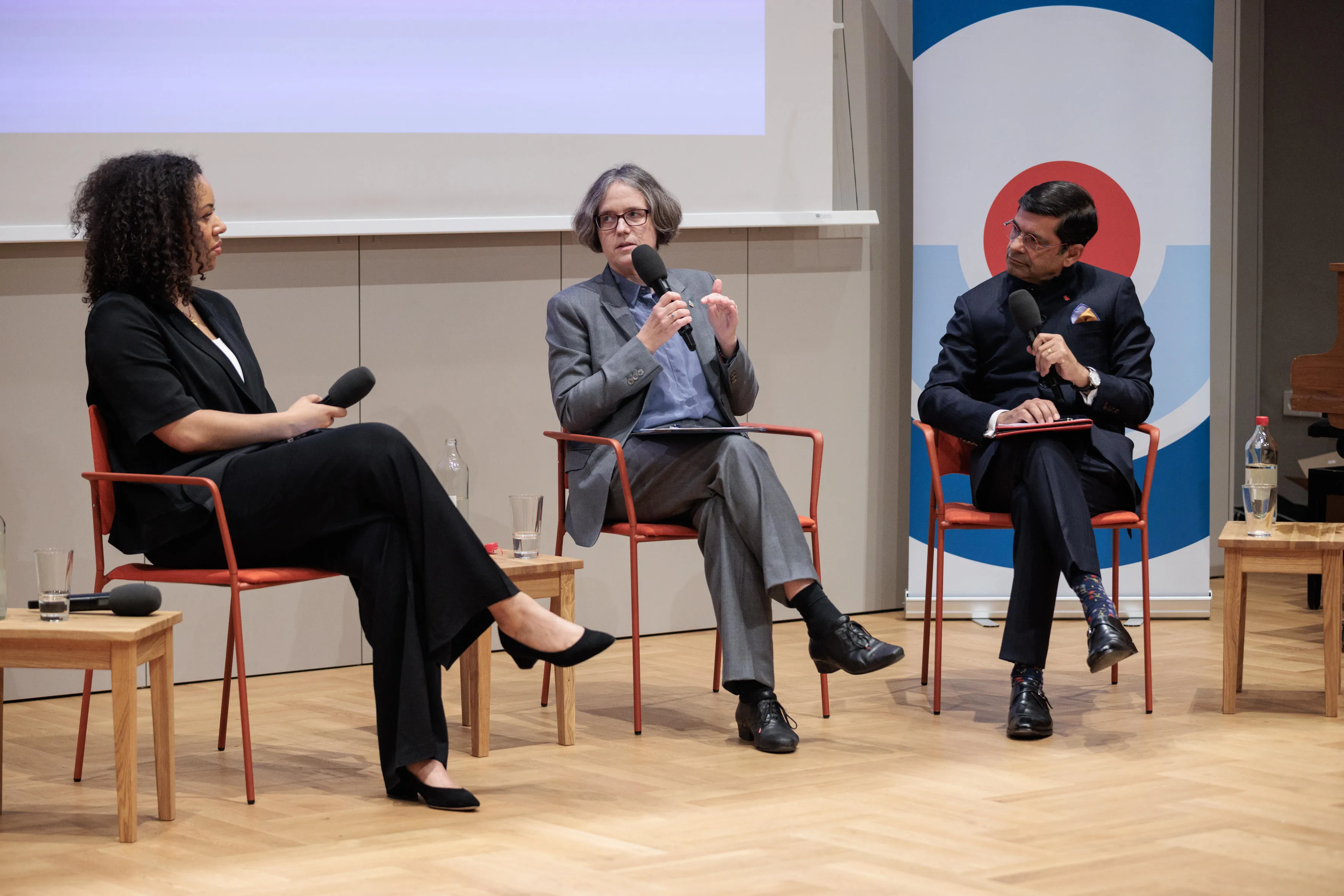 Three people—two in black suits and one in a grey suit—are sitting on a stage, engaged in conversation during an event.