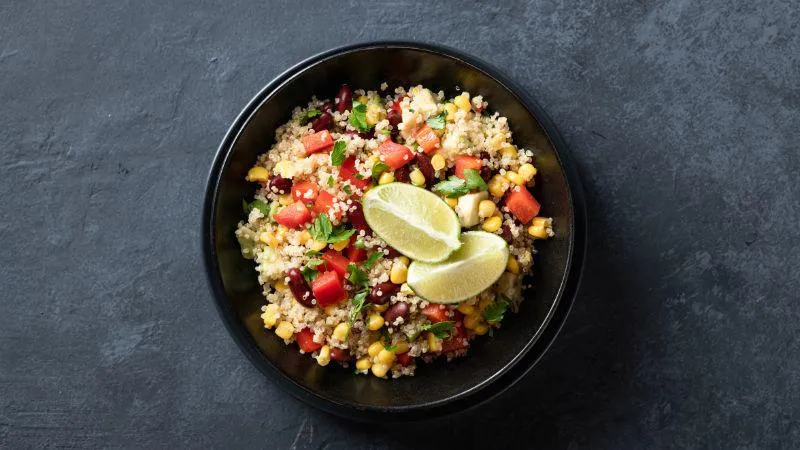 A black bowl filled with mixed quinoa salad, featuring beans, corn, chopped tomatoes, herbs, and two lime wedges on top, set on a dark textured surface.