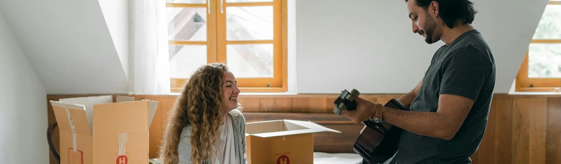Two flatmates moving into their new flat, there are boxes behind them and one is playing a guitar