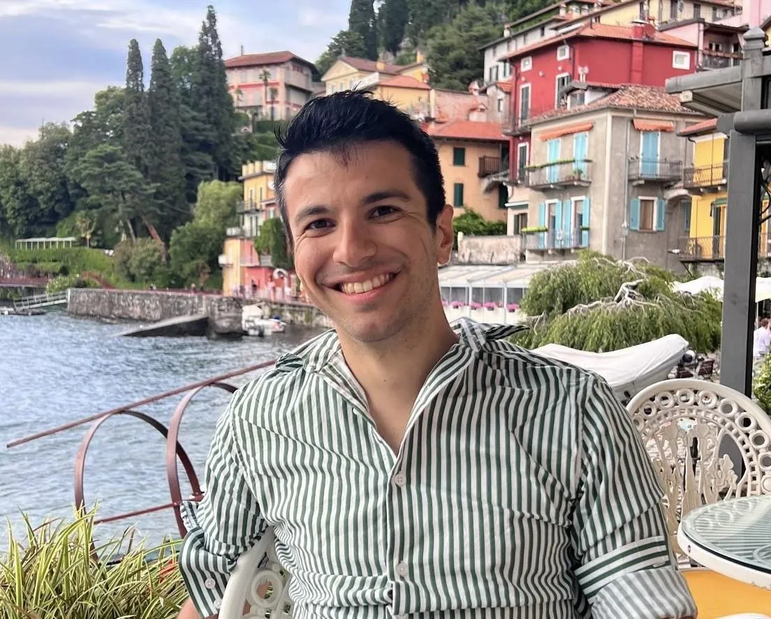 A man in a striped green and white shirt smiles on a terrace against a landscape of Italian coastal houses