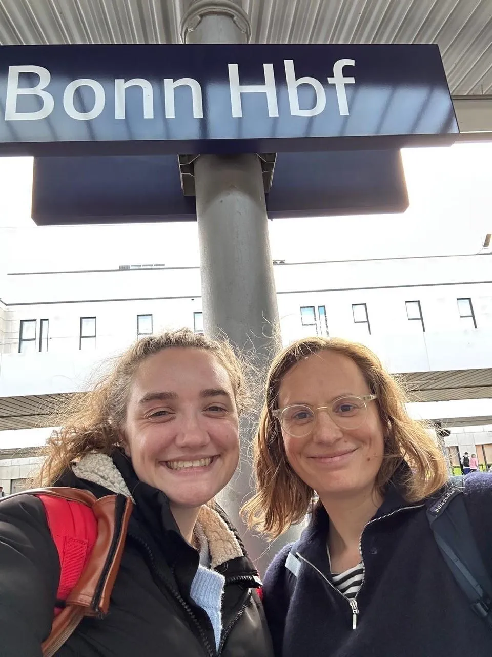 Alice and Aislinn smiling standing at Bonn train station under a blue station sign
