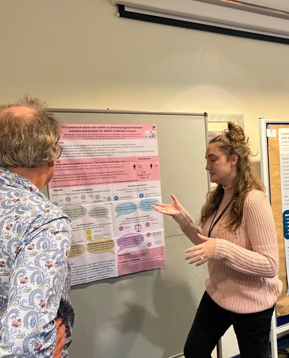 Alice presents a research poster to an attendee at a conference, gesturing as she explain key points