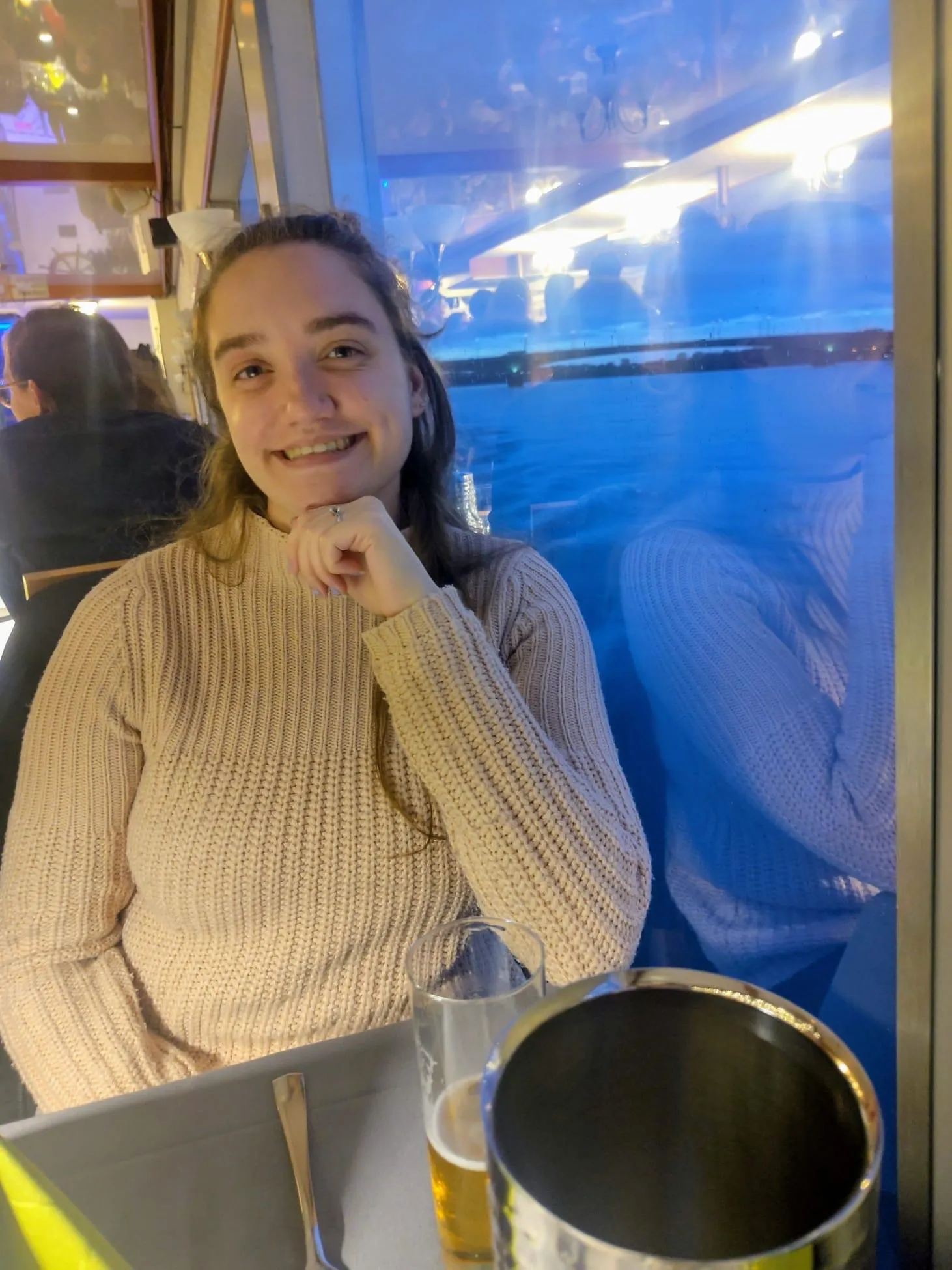 Alice smiles while seated at a restaurant table by a window in the evening, with a drink and cutlery in front of her