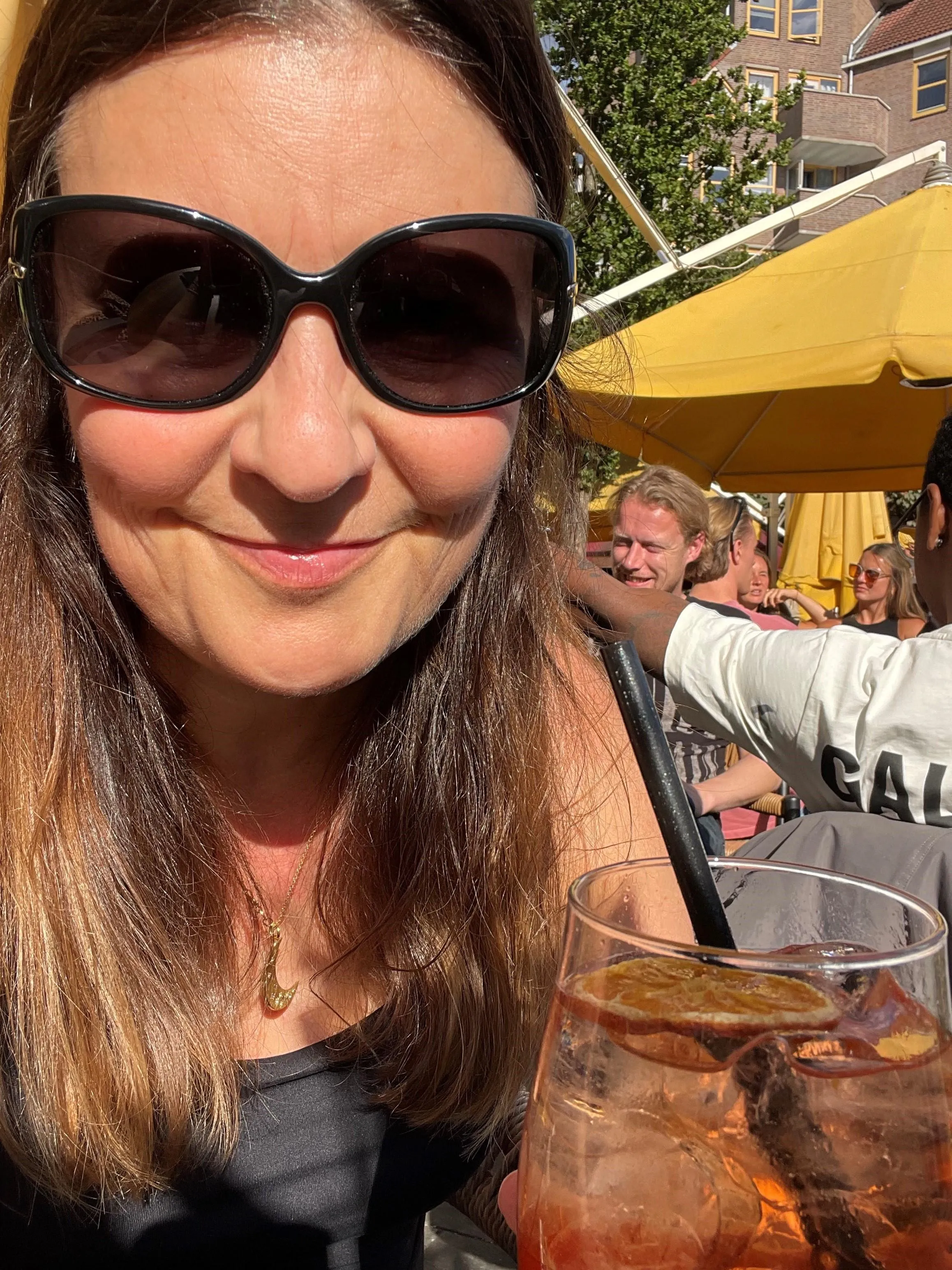 Angie in large black sunglasses smiling at an outdoor café, holding an orange iced drink