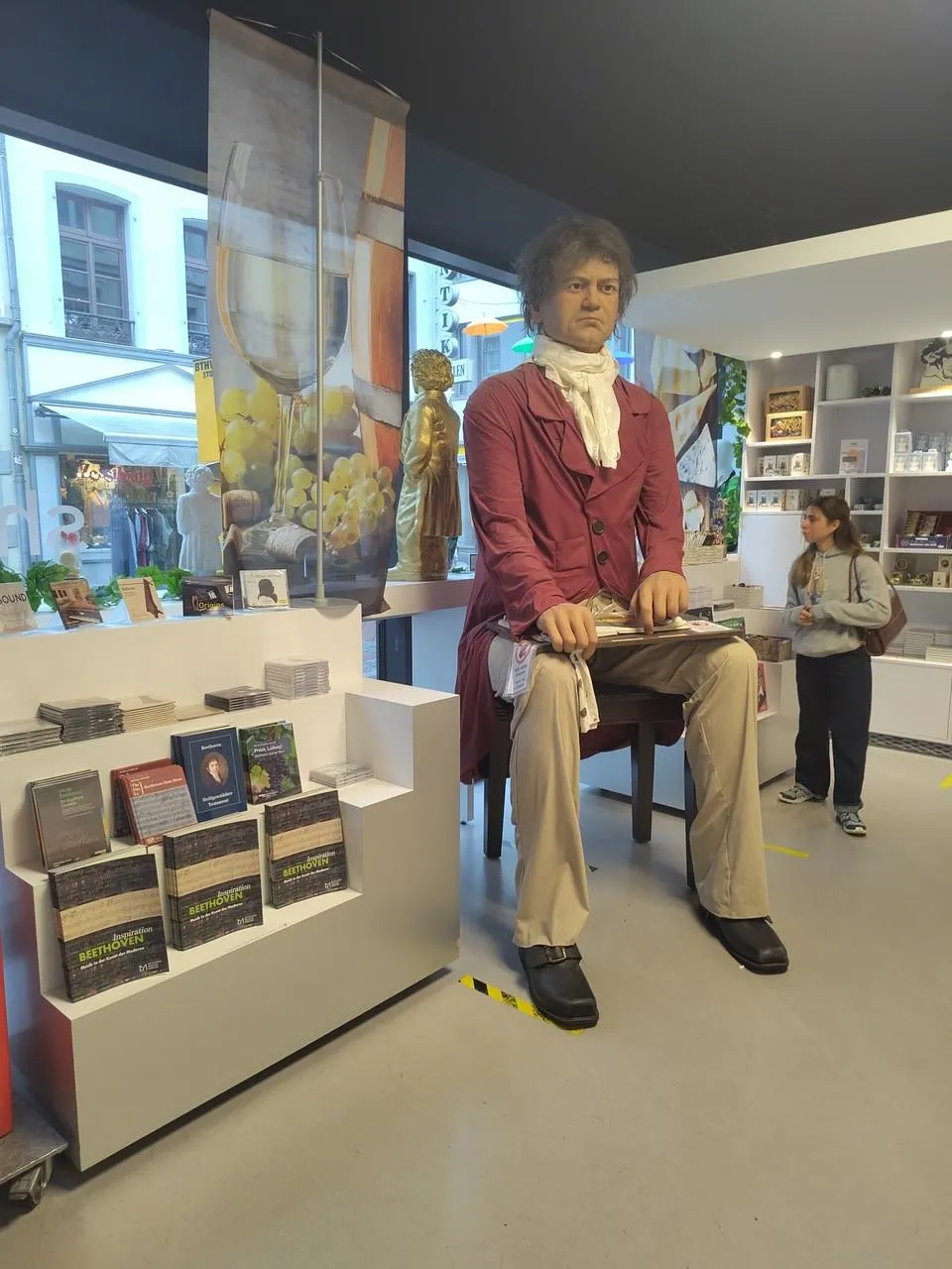 A large seated statue of Beethoven is displayed in a museum shop surrounded by CDs, books, and visitors