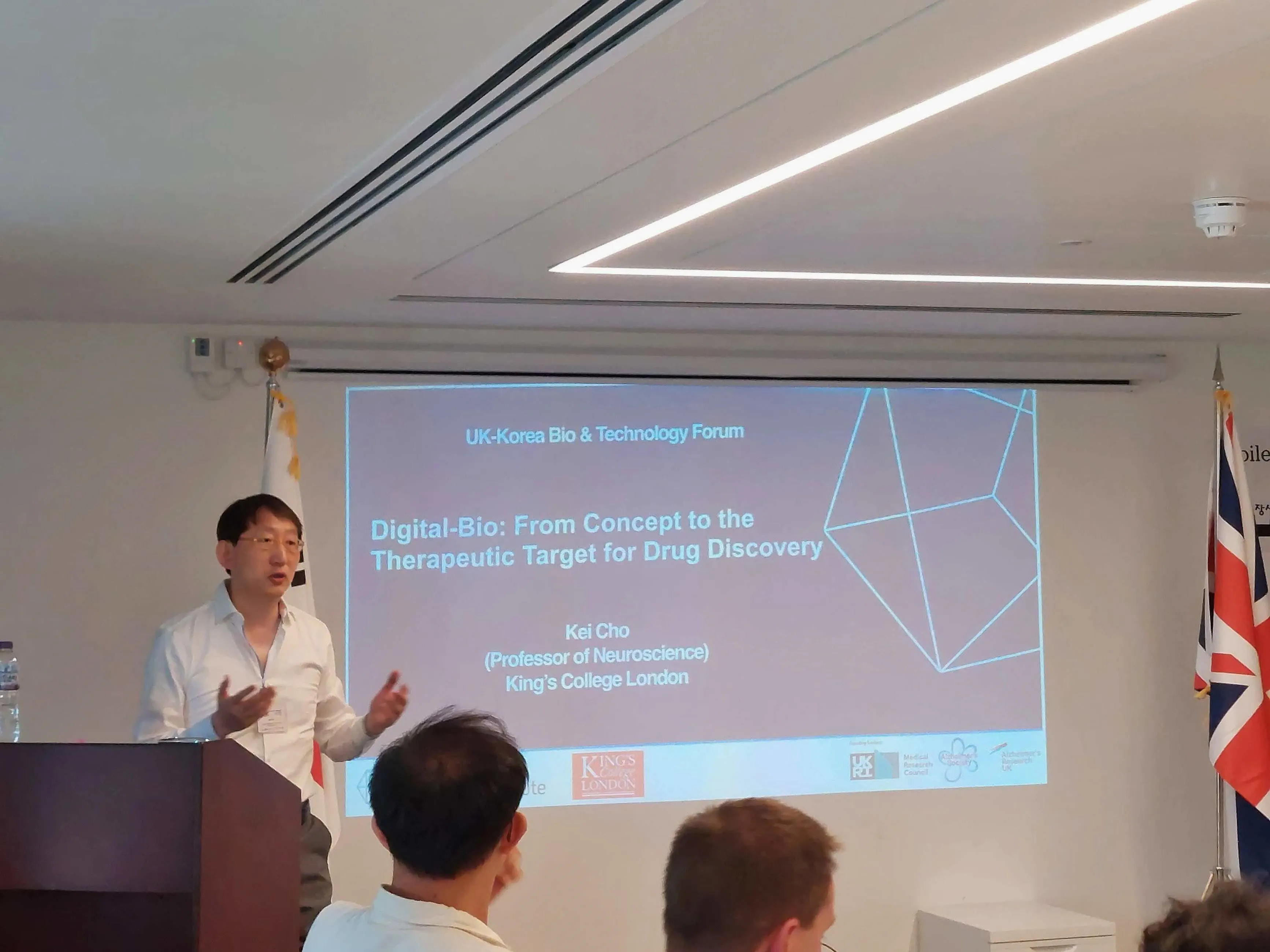 Professor Kei Cho standing next to his slide at the UK-Korea Bio&Tech Forum. Professor Cho wears glasses and a white shirt.