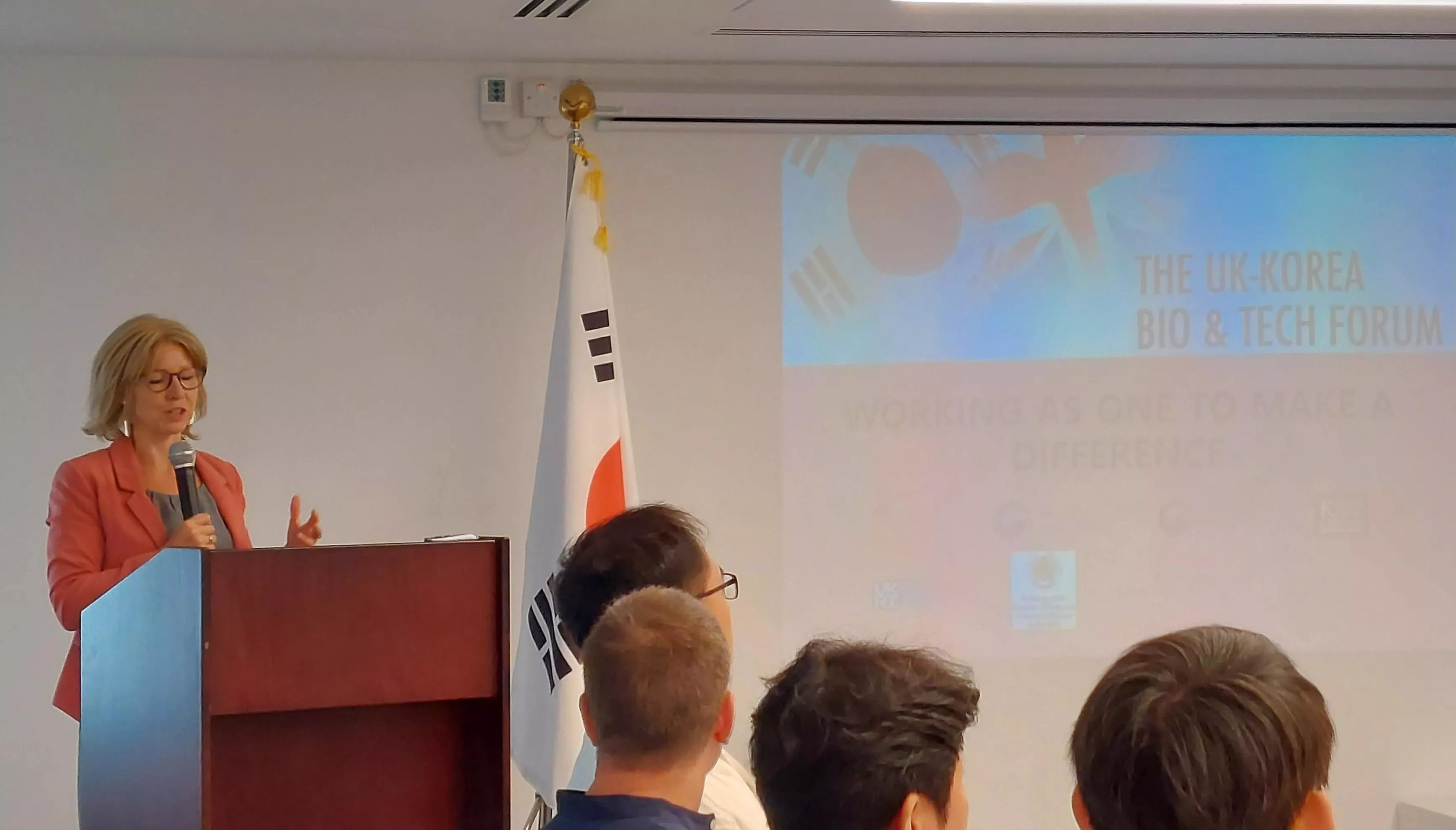 Ms Susie Kitchens stands behind a podium giving opening remarks to attendees of UK-Korea Bio&Tech Forum. She wears glasses, coral blazer, grey top and holding a microphone.