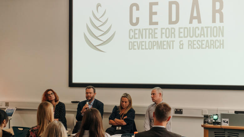 Four people seated at the front of a full seminar room. Screen behind says "CEDAR Centre for Education Development and Research"