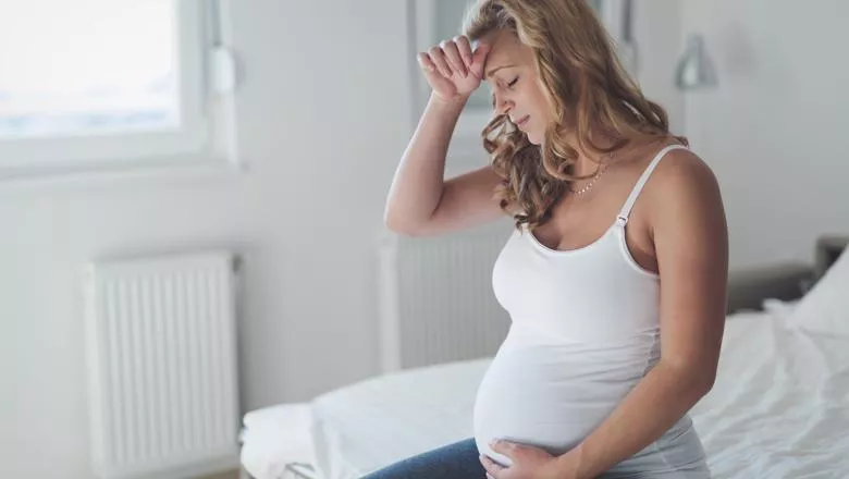 Pregnant woman sat on a bed resting her hand on her head looking distressed