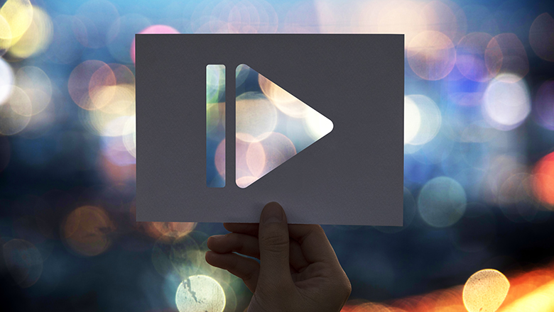 A hand holding up a piece of paper with a play button cut out, showing blurred lights behind