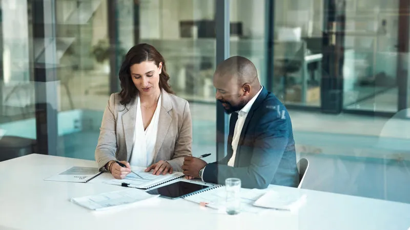 Two professionals sit at a white table in an office, discussing documents. The woman on the left is holding a pen over papers, while the man on the right gestures towards the documents. Both are dressed in business attire, with glass walls in the backgrou