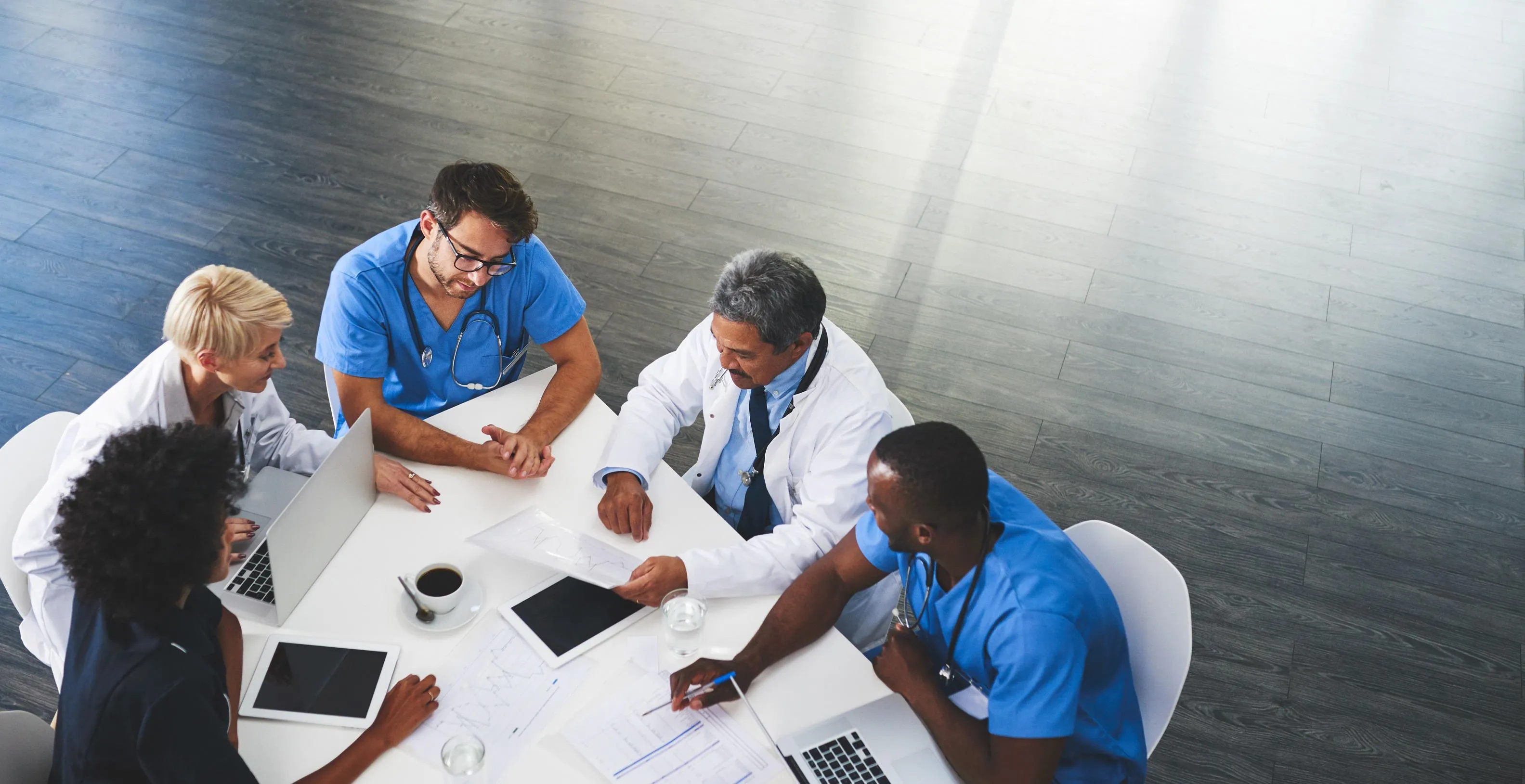 A group of medical professionals in scrubs and lab coats sit around a table, discussing documents and using laptops and a tablet. They appear engaged in a collaborative meeting, with a coffee cup and papers spread out on the table.