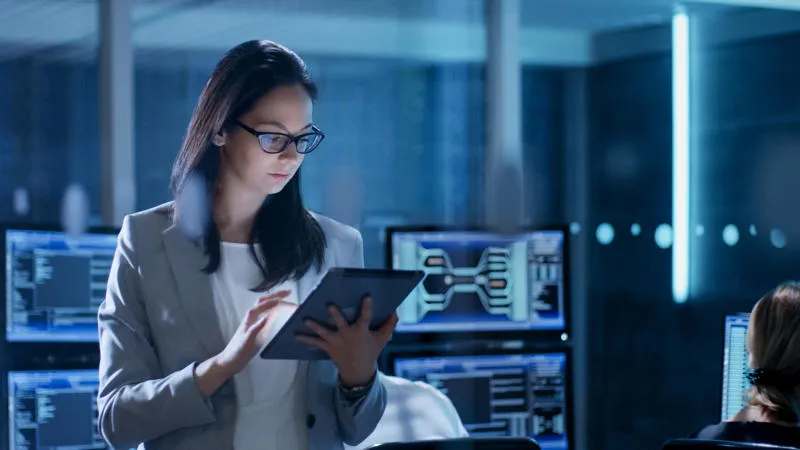 A woman in a business suit and glasses works on a tablet in a modern office environment, surrounded by computer screens displaying complex data and graphs. The room has a high-tech and focused atmosphere.