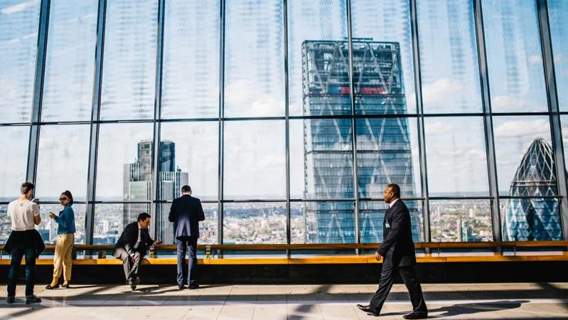 Man walking on pavement near people standing and sitting beside a curtain-wall building