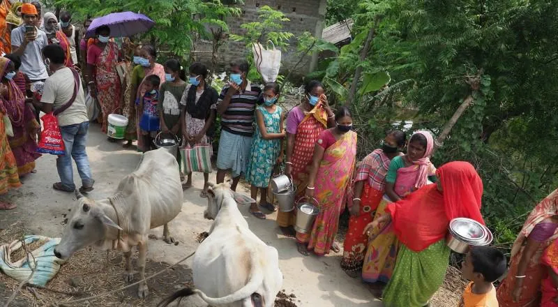 A line of people in brightly-coloured clothes queue for food with pans.