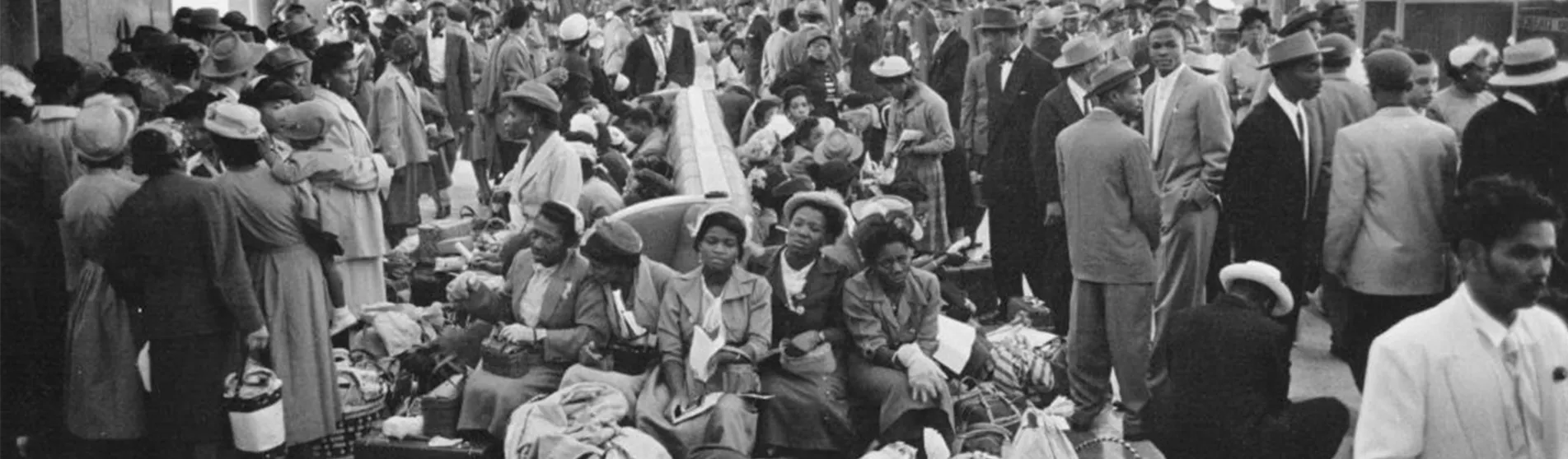 A large group of people likely to be a cohort of people aboard on Windrush are waiting. The picture is in black and white.
