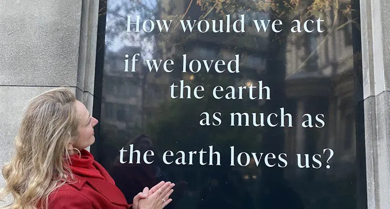 A woman in a read coat is tilting her head towards a billboard with writing on it