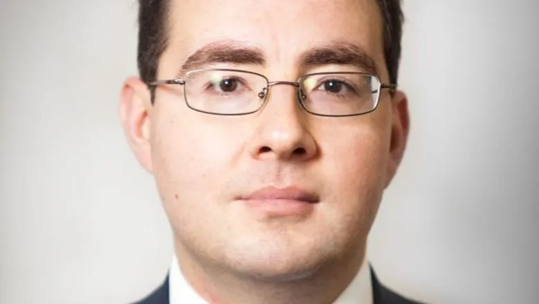 A man with short dark hair and glasses is in front of a white background