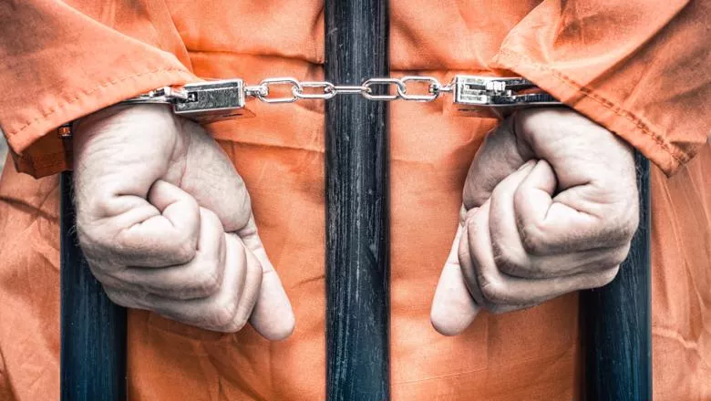 A man in orange overalls has his hands cuffed around prison bars